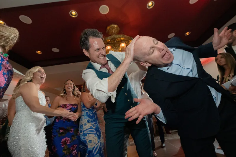 Two men dance together playfully, one tilted back dramatically, while guests watch at the reception.