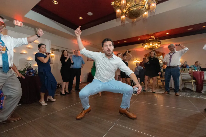 Rhett performs an energetic squat-jump dance move on the floor surrounded by applauding guests.