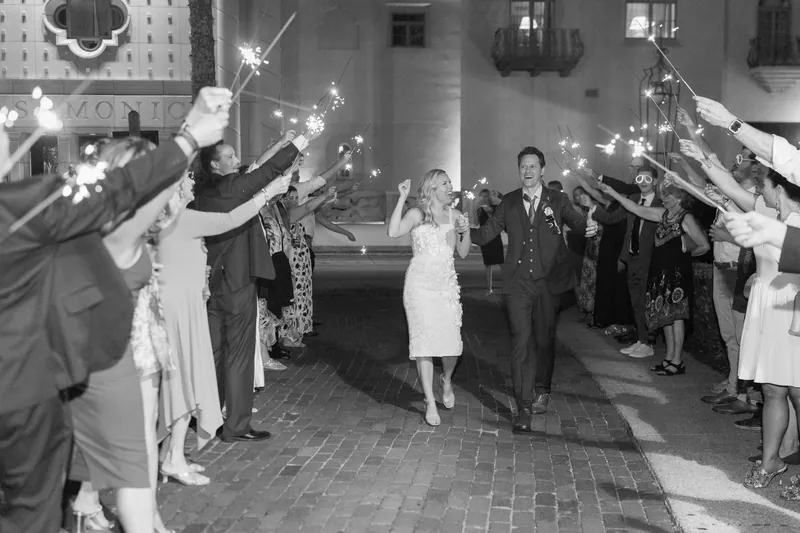 Guests line a columned courtyard holding sparklers aloft as Kerry and Rhett walk between them in black and white.