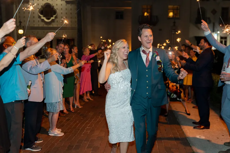 Kerry in a white sequined dress and Rhett in a dark suit walk through a tunnel of guests holding sparklers
