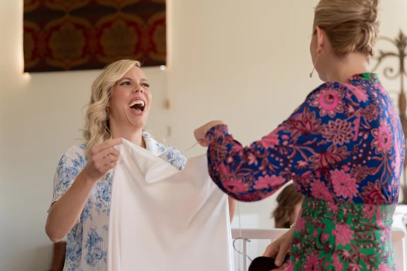 Kerry laughs while receiving a white fabric from a companion in a matching floral dress.