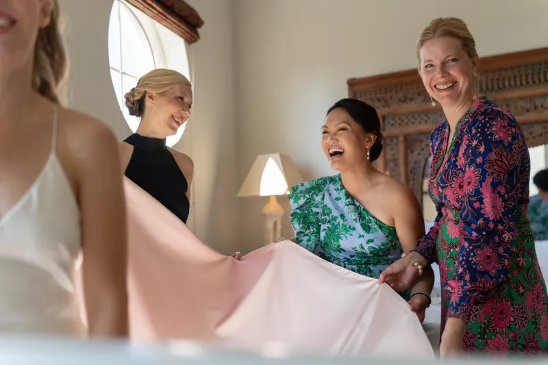 Three women smile while holding up Kerry's pink wedding dress in a hotel room.