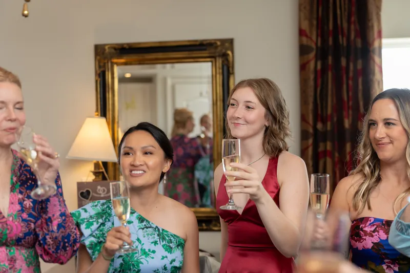 Four women in festive attire raise champagne glasses indoors, reflected in a gold-framed mirror behind them.