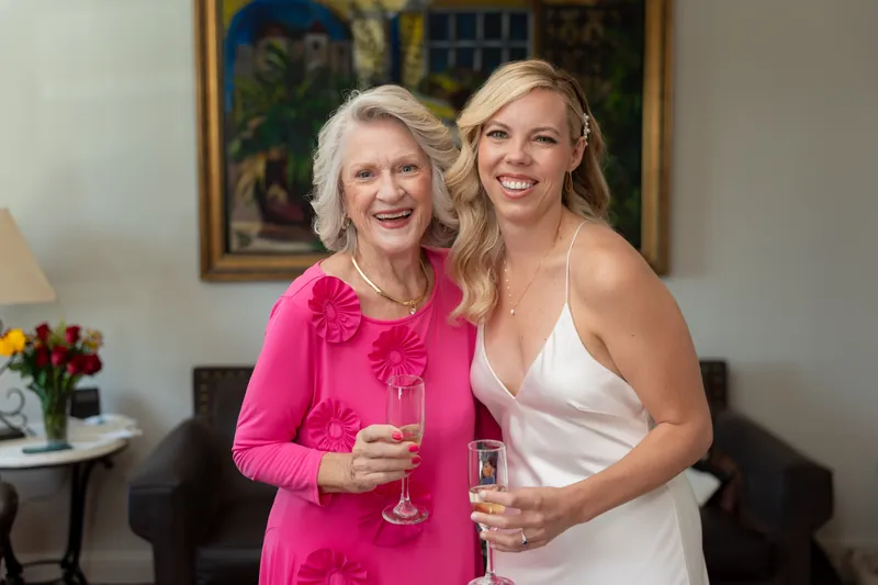 An older woman in hot pink stands beside Kerry in white, both holding champagne flutes and smiling at Casa Monica Resort &