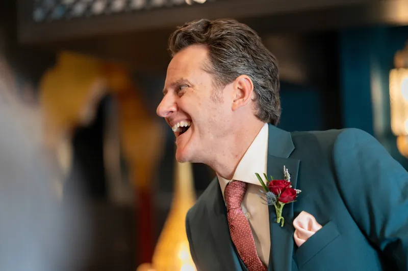 Rhett laughs heartily in a dark suit with a burgundy tie and rose boutonniere during the celebration.