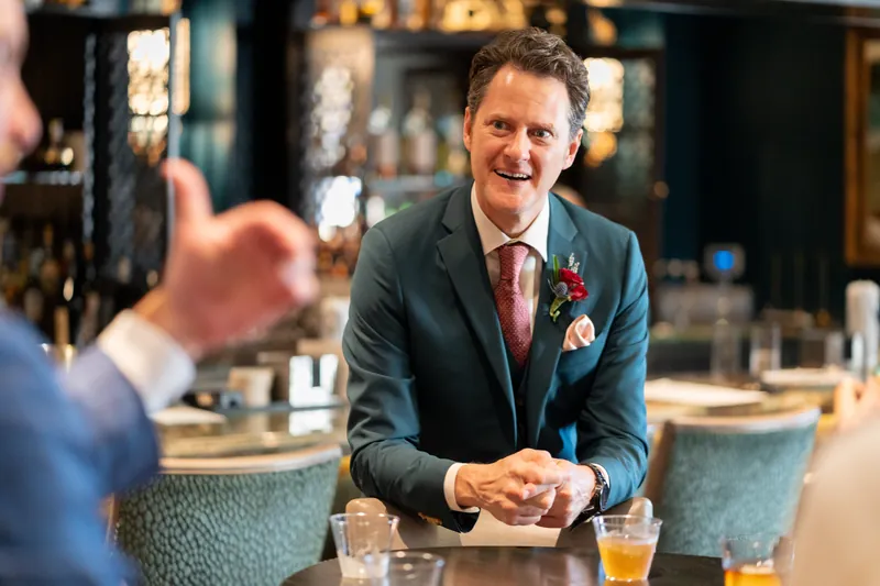 Rhett smiles while seated at a bar, wearing a dark blazer and burgundy tie with a red boutonniere.