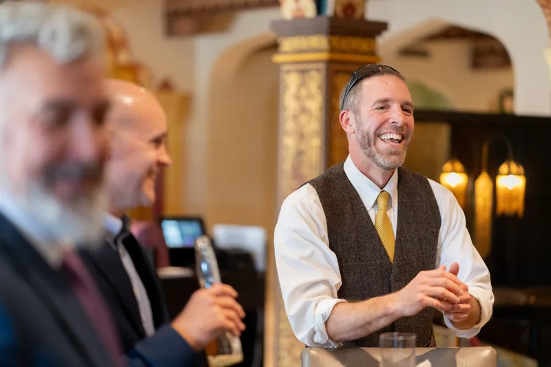 A man in a brown vest and white shirt gestures while speaking to guests in the elegant venue.