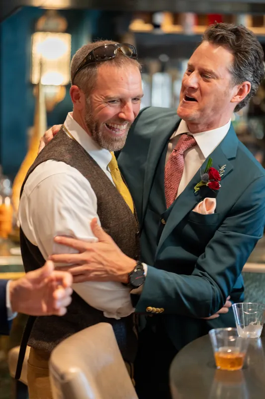 Two men embrace and laugh together during the reception, both wearing formal attire with boutonnieres.