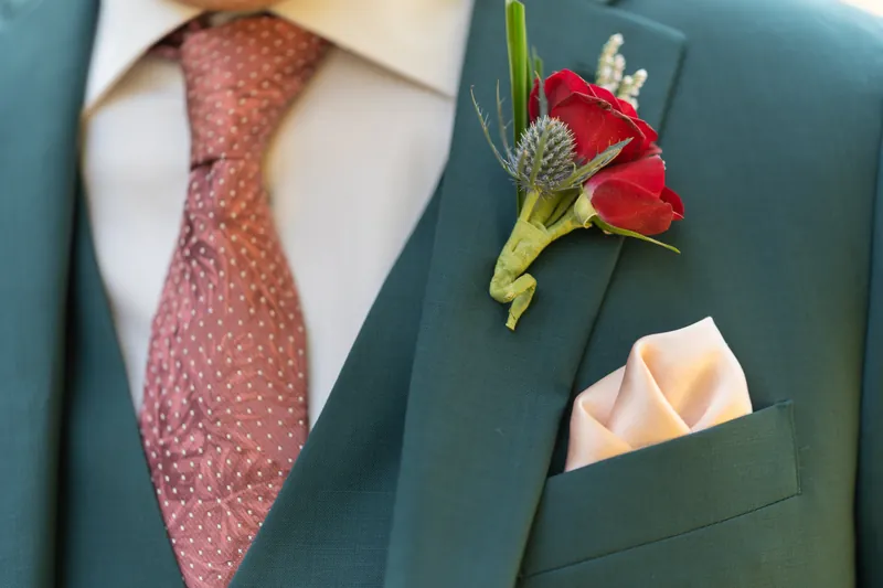 Close-up of a teal blazer with a burgundy tie and red carnation boutonniere paired with a cream pocket square.