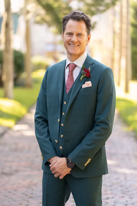 Rhett stands in a teal suit with burgundy tie and boutonniere on a brick pathway at Casa Monica Resort & Spa.