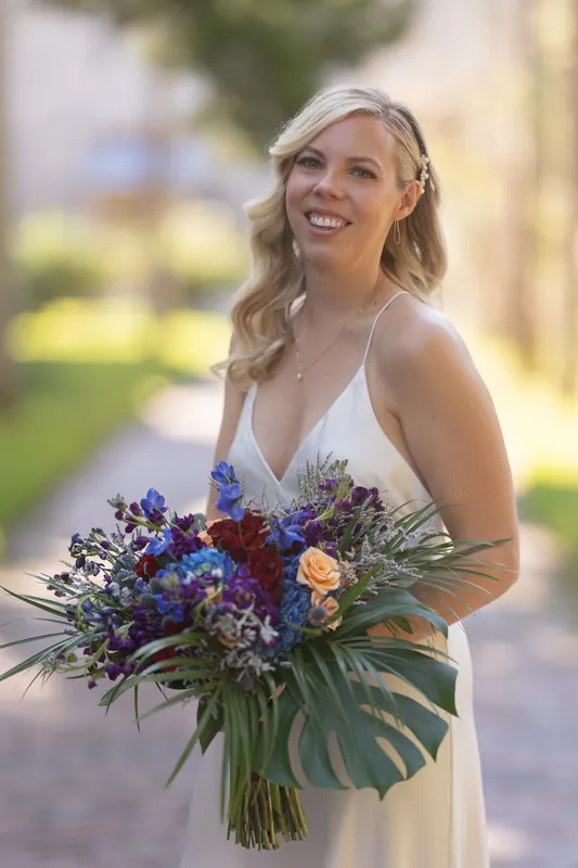Kerry holds a lush bouquet of deep blue, burgundy, and peach flowers with monstera leaves at Casa Monica Resort & Spa.