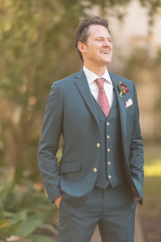 Rhett smiles in a slate-blue suit with burgundy tie and small red boutonniere, positioned outdoors with soft-focus greenery