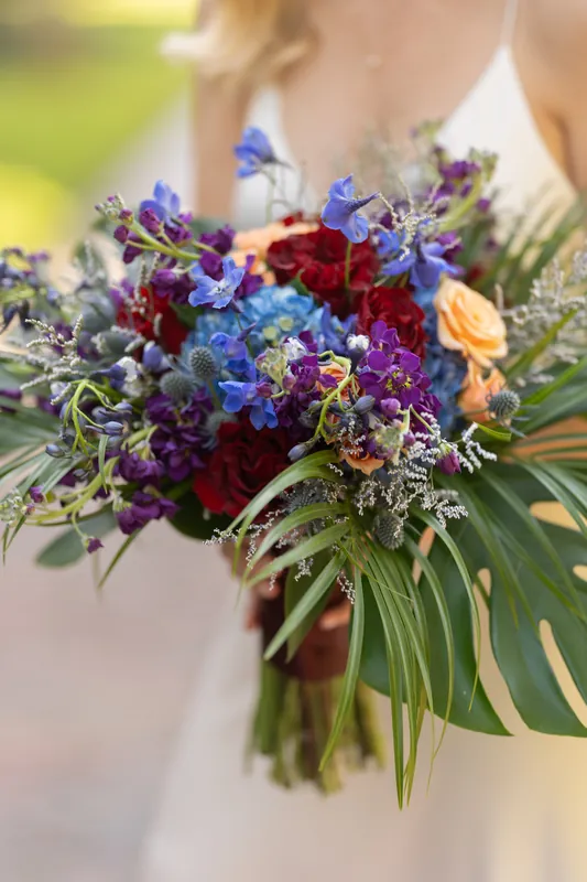 Dense arrangement of purple, blue, red, and peach blooms with green monstera fronds fills the frame in Kerry's wedding