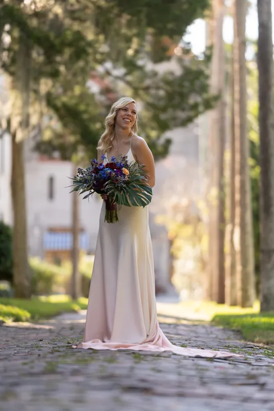 Kerry stands centered on a stone pathway lined with tall cypress trees, holding her vibrant bouquet in her cream wedding