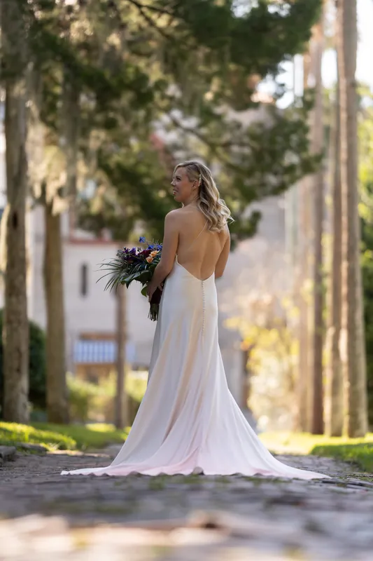 Kerry stands alone on a tree-lined pathway in a flowing white gown with an open back, holding a dark bouquet and looking