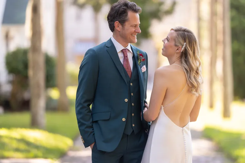 Rhett in a navy double-breasted suit and Kerry in her backless white dress walk together down a shaded arcade, smiling