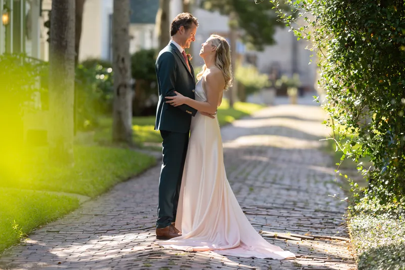 Kerry and Rhett embrace on the brick pathway, framed by green hedges and columned architecture on either side.