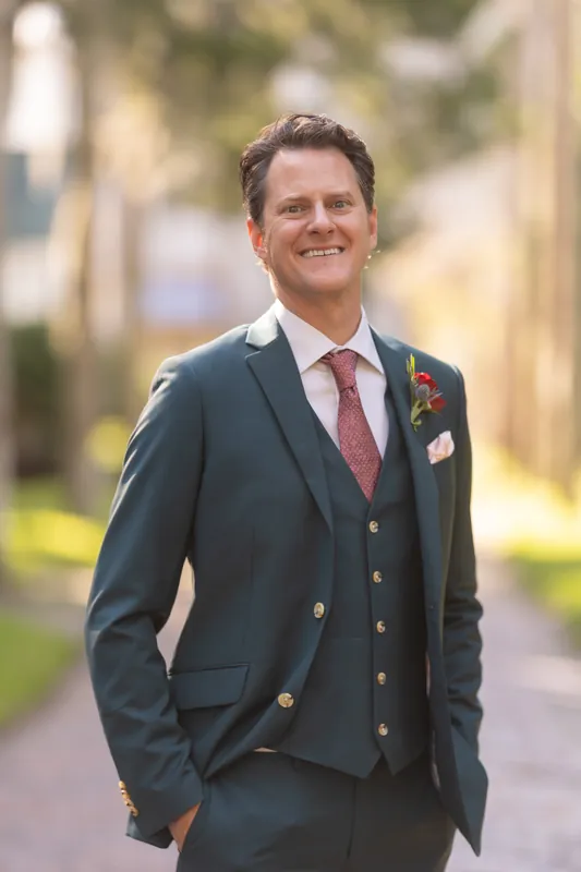 Rhett stands alone on a shaded brick path wearing a navy double-breasted suit and burgundy tie with a boutonniere.