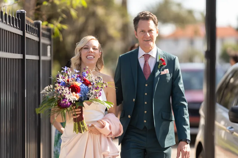 Kerry and Rhett walk together along a fence-lined street, Kerry holding a colorful mixed bouquet and Rhett in his navy suit.