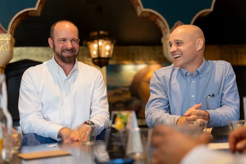 Two men in light blue and white shirts sit at a table indoors, conversing and smiling during the reception.