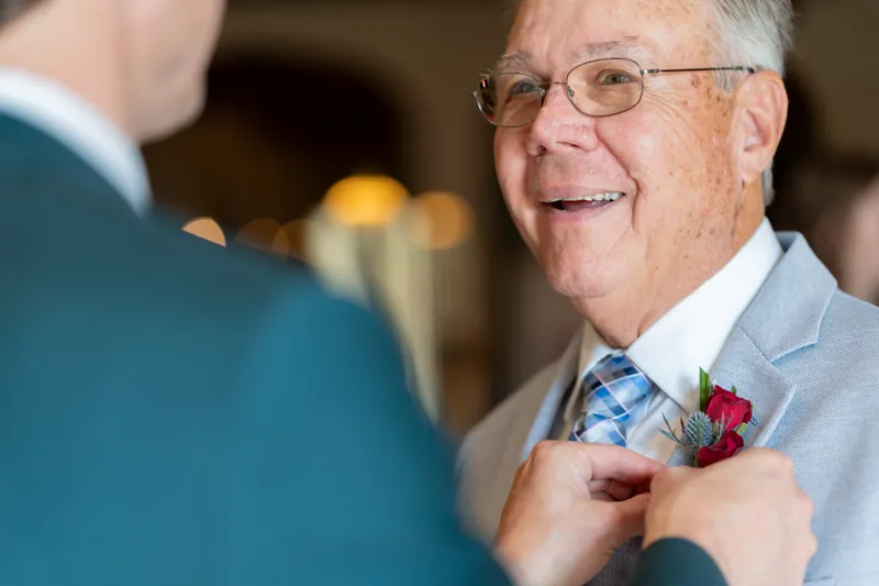 A man in a light blue suit adjusts a boutonniere on someone's lapel during wedding preparations.