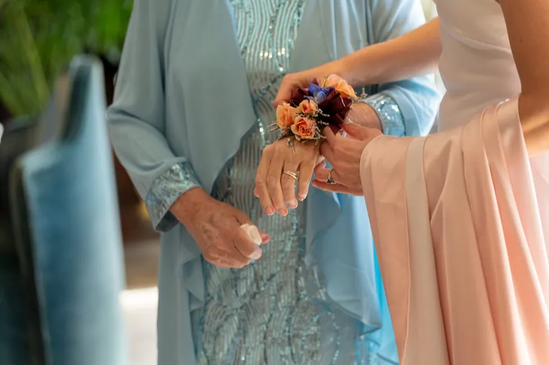 Hands exchange a small orange and burgundy bouquet between two people in formal attire.