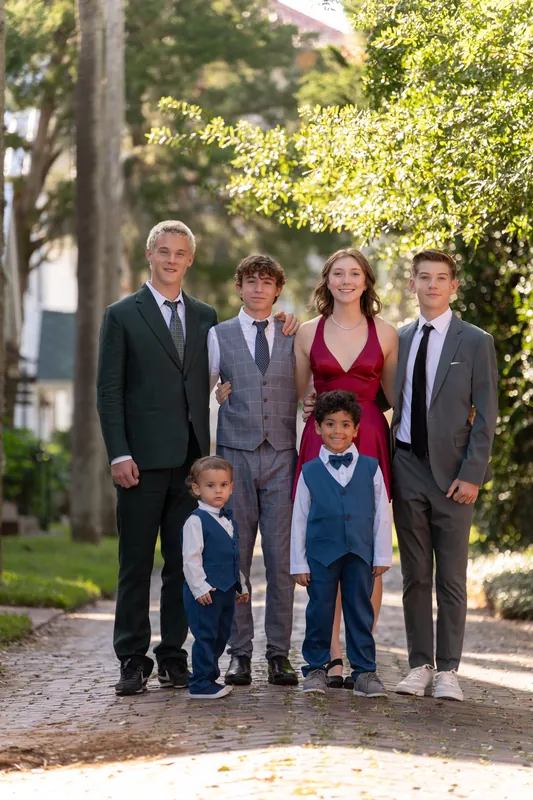 Five family members and two young children pose together on a brick pathway lined with trees.