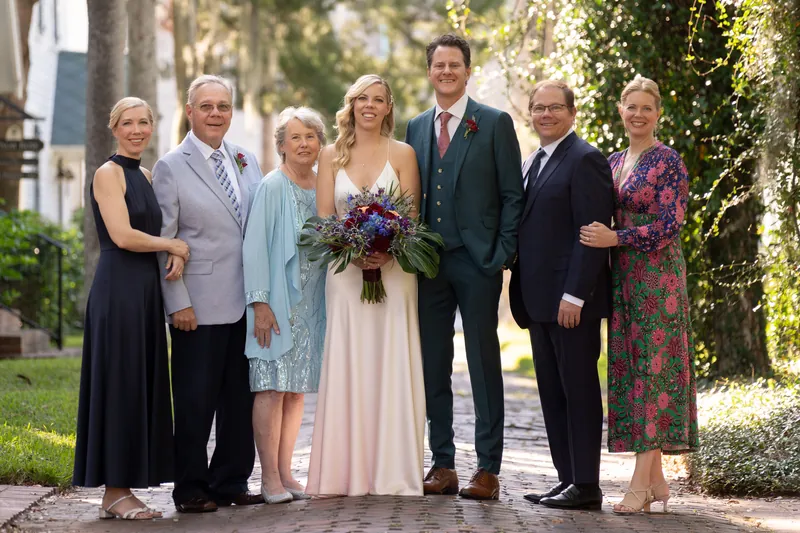 Seven family members stand together on a shaded brick pathway, with the center couple holding a burgundy bouquet.