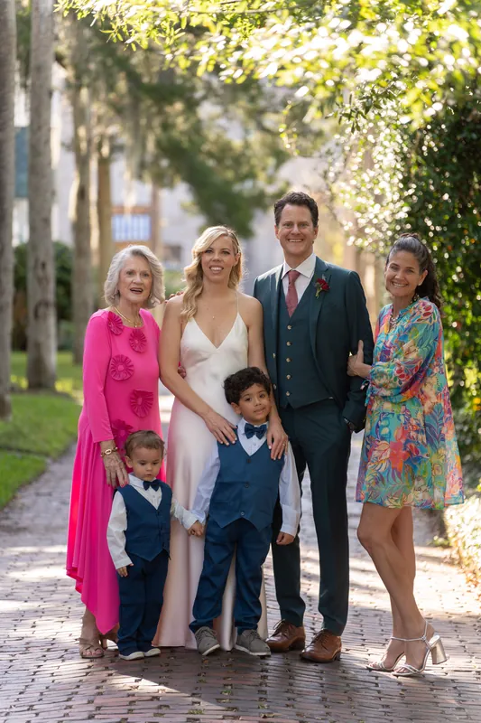 A family of five, including two children in blue vests, stands on a brick path with flowering gardens behind them.