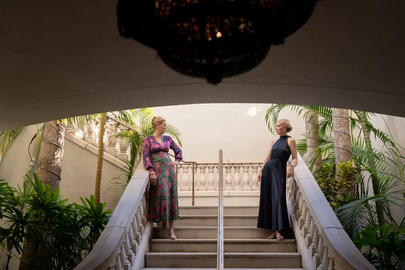 Two people ascend a grand staircase with white railings beneath an ornate chandelier at Casa Monica Resort & Spa.