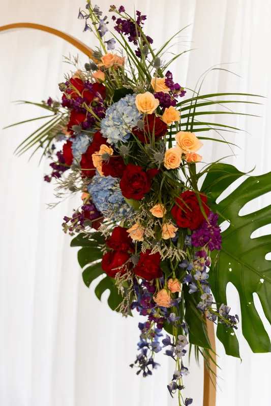 Lush bridal bouquet featuring deep red roses, burgundy dahlias, orange ranunculus, and trailing vines arranged vertically.