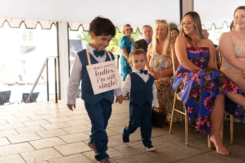 Two young children walk down the aisle holding hands while guests smile from their seats at the indoor ceremony.