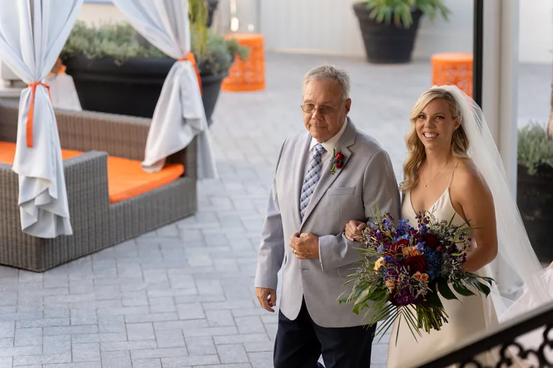 Kerry walks arm-in-arm with a companion down the aisle, carrying a deep jewel-toned bouquet at the altar.