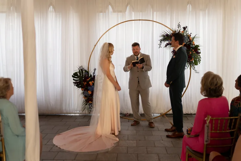Kerry stands in a flowing white gown beside Rhett in a tan suit as an officiant speaks under a floral arch during
