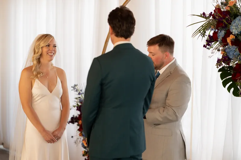 Kerry smiles at Rhett during their ceremony as the officiant looks down at notes between them.