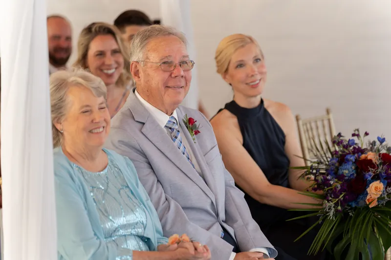 Kerry's parents and family members sit together watching the ceremony, with dark and coral flowers visible nearby.