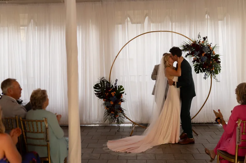 Kerry and Rhett kiss under a circular arch decorated with dark greenery while guests applaud around them.