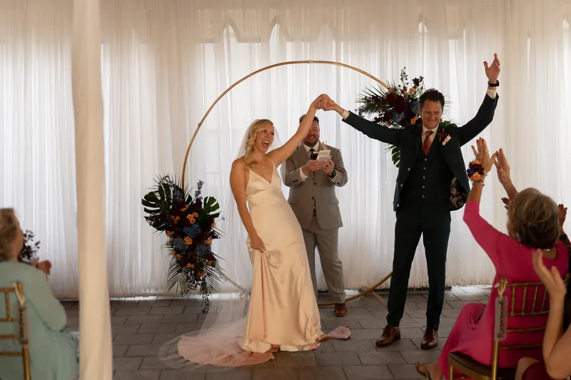 Kerry and Rhett celebrate with raised arms as guests cheer, standing beneath the decorated arch with sheer white draping