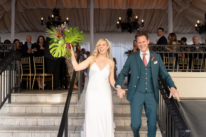 Kerry and Rhett walk down outdoor steps hand in hand, Kerry holding her bouquet high as they exit their reception.
