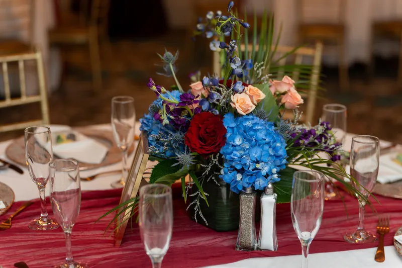 Lush centerpiece of blue hydrangeas, red roses, and peach flowers sits on burgundy linens with place settings and glassware.
