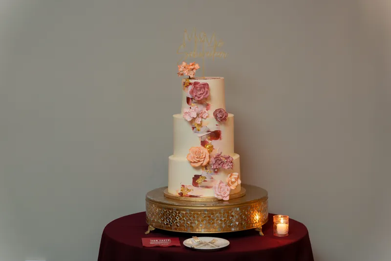 Three-tiered wedding cake with cream frosting and coral floral accents sits on gold base with burgundy linens