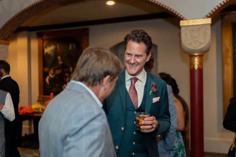 Rhett in a teal blazer and pink tie converses with a guest in a light blue shirt during the reception.