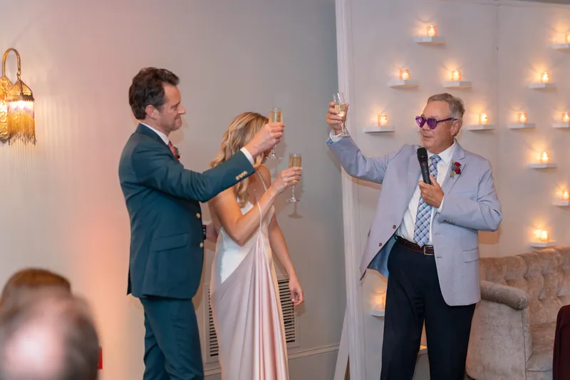 Rhett toasts alongside two companions, all three raising glasses indoors beneath warm lighting.