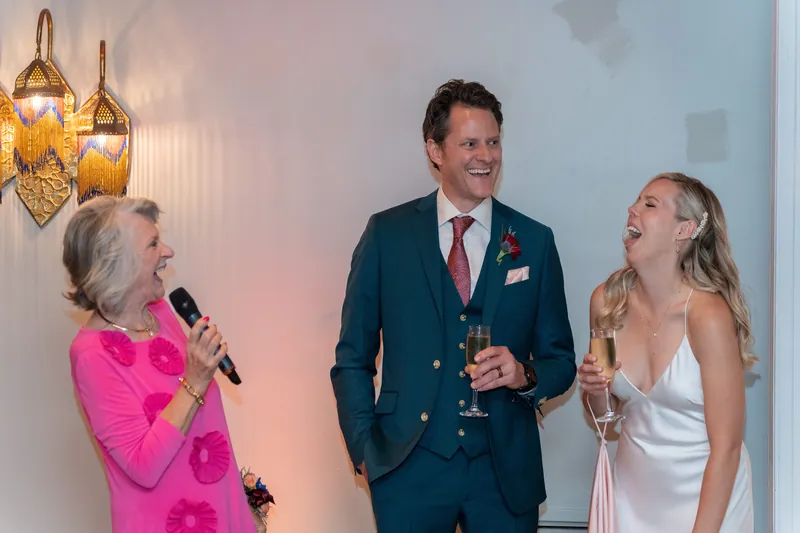 A woman in a bright pink jacket speaks into a microphone while Rhett in a navy blazer and Kerry in a white sleeveless dress