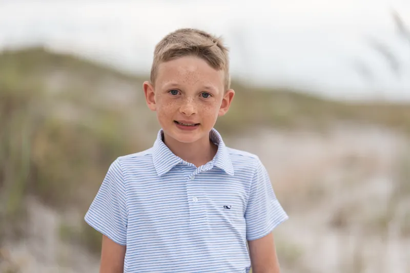 A young child in a light blue striped polo smiles at the camera with sand dunes behind them at Vilano Beach.