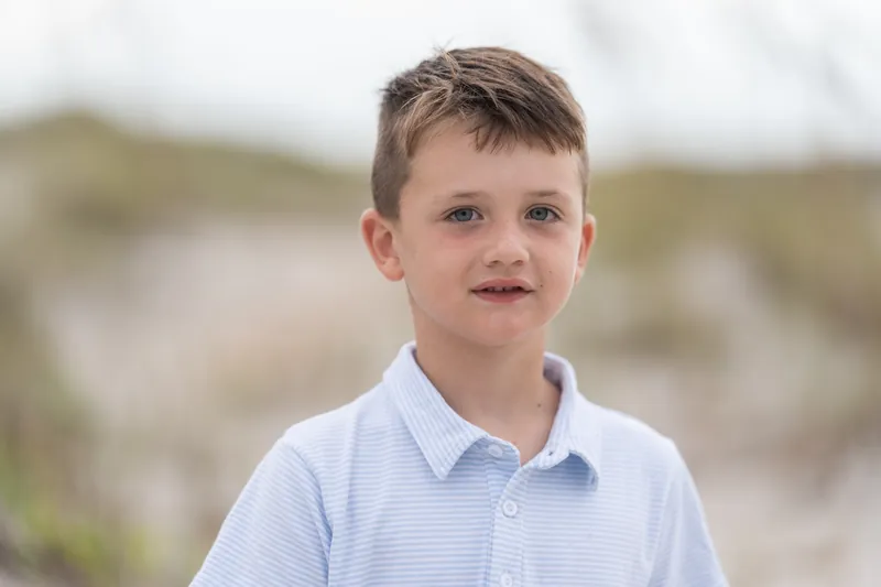 A child wearing a pale blue button-up shirt stands on the sand with beach grasses and dunes softly blurred in the background.