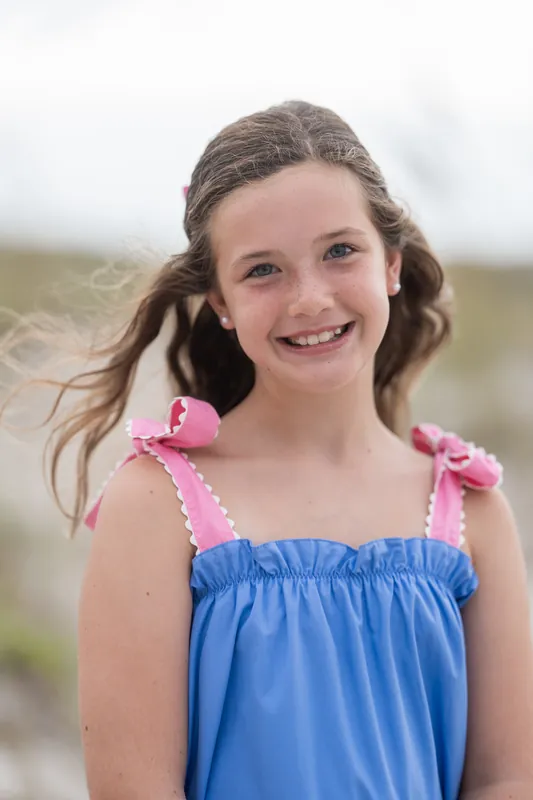 A young person in a blue sundress with pink ribbon straps smiles with tousled hair, beach dunes visible behind them.