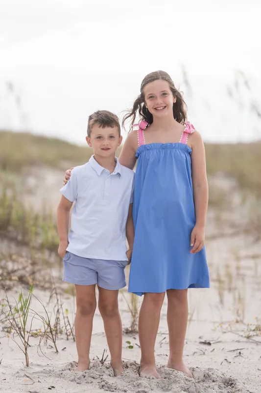 Two children stand together on the sand at Vilano Beach—one in a light blue polo and shorts, the other in a blue dress with pink accents.