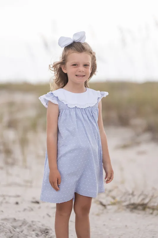 A child in a lavender dress with ruffled sleeves and a white bow stands barefoot on the sand with beach vegetation behind them.