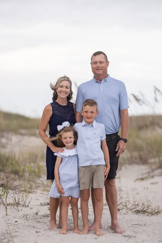 A family of four stands barefoot together on the sand at Vilano Beach, with dunes and coastal grasses in the background.